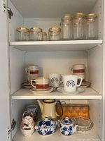 Stoneware mugs in red stripe and floral patterns, matching saucers, a ceramic cat-shaped napkin holder, yellow ceramic pitcher, blue and white ceramic lidded containers displayed on shelves inside white cabinet.