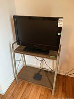 Full view of the Emerson flat screen TV positioned on a metal frame stand with two wooden shelves, placed against a white wall on wooden floor.