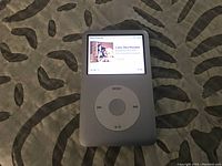 Front view of iPod Classic showing color display with song playing and white click wheel.