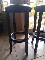 Side view of one bar stool showing wooden legs, circular footrest, and the faux leather seat with some creasing.