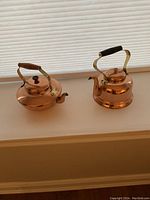 Front view of two copper kettles on a white surface near a window with blinds, showing brass handles and wood grips.