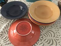 Set of six pasta bowls in various colors stacked and displayed on a table including bottom of one red-orange bowl showing markings.