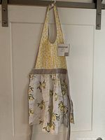 Apron hanging on white door showcasing yellow polka dots on top, lemon and leaf print on skirt portion, beige band, and button closure strap.