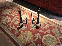 Two antique brass andirons standing on floral patterned carpet with sunlight casting shadows.