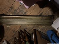 Single photo showing the full brass fireplace fender lying flat on wooden floorboards with surrounding miscellaneous items.