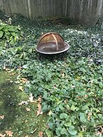 Copper fire bowl placed outdoors on ground with surrounding green plants. The bowl shows weathering and patina, with a dome mesh cover on top.