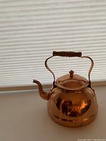 Copper kettle sitting on a surface showing the full kettle with copper body and wooden handle, reflecting lighting and surrounding environment.