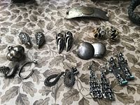 Top down view of various silver-tone clip-on earrings, rope-style earrings, and a metal hair clip with medallion detail on a floral fabric background.