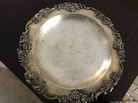 Round silverplate on copper tray with ornate grapevine decoration on scalloped rim and central engraved monogram. Shows signs of age and tarnish.