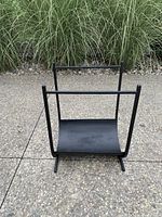Front view of a black matte finish wrought iron log holder placed on ground outdoor pavement with green grass background.