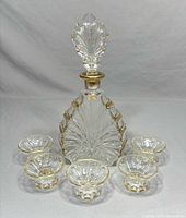 Front view of crystal decanter with stopper and five matching cups arranged around it, showing gold accent details and starburst cut pattern.