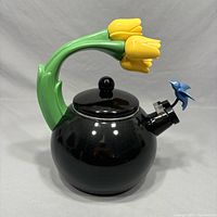 Front view of black enamel teapot with green tulip-stem handle, yellow tulip flowers, and blue hummingbird whistle attached to the spout.
