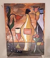 Front view of the acrylic painting showing the colorful still life composition with bottles, glass, flowers, grapes, and fruit bowl. Artist signature visible lower left.