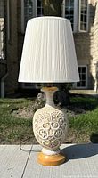 Front view of mid century ceramic table lamp outdoors showing intricate ceramic body, wooden base, and tall pleated fabric shade.