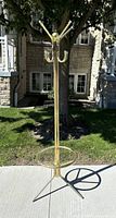 Full view of vintage brass tone coat and umbrella rack standing outdoors on sidewalk with tree and house in background. Eight hooks visible with umbrella ring at base.