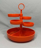 Front view of the orange plastic snack tower showing the ring handle, three small circular bowls on top and the segmented larger base tray.