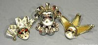 Three vintage Italian Venetian carnival masks arranged together on a grey background. Each mask features detailed painted faces with decorative elements including ribbons, bells, and pearls.