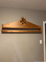 Photo of a wall-mounted wooden blanket rack and shelf in a medium wood tone with a decorative carved ribbon on the triangular top edge.