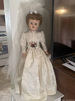 Front view of bride doll dressed in ivory patterned wedding gown with puff sleeves and floral belt, long white veil flowing down back.
