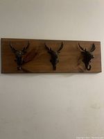 Wooden plank mounted on a wall with three metal animal head-shaped hooks with antlers.
