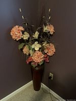 Tall artificial floral arrangement placed in a corner next to the wall with an electrical outlet visible near the base. The flowers are peach-colored hydrangeas and cream lilies with greenery in a tall brown vase.