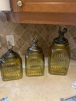 Set of three ribbed amber-colored glass canisters with metal lids topped with rooster finials arranged on a kitchen counter.