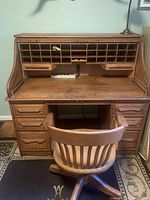 Front view of the roll top desk with the top open showing pigeonhole compartments and drawers; the wooden swivel chair is positioned in front.