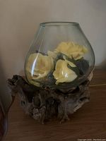 Clear glass bowl filled with yellow artificial roses and green leaves, resting on a natural wood base, shown against a plain background.