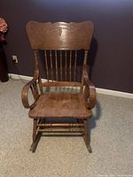 Front facing image of wooden rocking chair showing carved backrest, spindle back supports, curved armrests, and rocker legs.