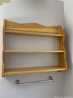 Wooden wall shelf with three flat shelves and decorative curved side panels, mounted on a white wall above two towel bars.