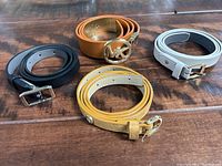 Four belts laid out on wood surface showing different colors and buckle styles including Michael Kors belt.