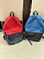 Front view of two youth Champion backpacks side by side, one red with black pocket and bottom, one blue with black pocket and bottom, showing zippers and logo patches.