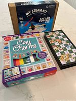 Photo shows the three main items of the lot: DIY STEAM kit box labeled 'Easy Air Powered Car', a Klutz Clay Charms kit with colorful modeling clay and instruction book, and a foldable travel-sized Snakes and Ladders board game closed case.