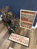 Two festive twig deer sculptures with pine branch and ornament accents beside two framed Christmas-themed wall hangings on a wood floor against a navy blue wall.