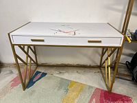 White desk with a gold metal geometric leg frame, showing paint marks on the desktop and a drawer with gold handles.