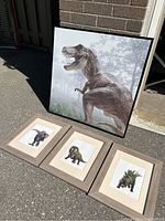 Large framed canvas print of T-Rex and three smaller framed dinosaur illustrations on ground outdoors
