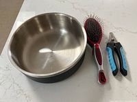 Stainless steel dog bowl with black bottom, dog grooming brush with red tips on metal bristles, and blue nail trimmers with black grip.