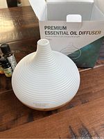 Photo of white ribbed essential oil diffuser, wood-grain base, packaging box, and some essential oils nearby.