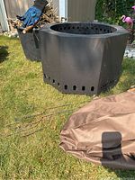 Full view of the black octagonal metal fire pit with ventilation holes, blue fireproof gloves resting on firewood container, and folded brown rain cover on grass.