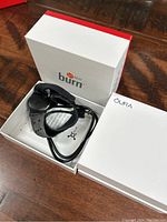 Orange Theory heart monitor device with strap and charging cable inside open branded box, alongside closed Oura box.