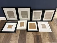 Overview of 9 picture frames in black and white laid on wooden floor against blue wall.