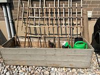 Photo shows raised wooden garden bed with five wooden trellises against a brick wall, green watering can, some vine clips, and a roll of burlap fabric inside the bed.