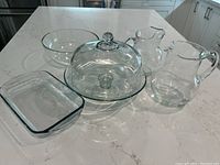 Set of glass kitchenware items displayed on counter: cake stand with dome lid, two pitchers, large serving bowl, rectangular casserole dish