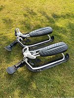 Two black and white GT Stiga Pro snow racers, each with black skis and pedals, white steering columns, and black padded seats laid side by side on grass.