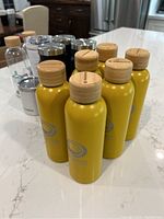 Ten yellow insulated water bottles with wooden caps, two transparent uninsulated water bottles in background