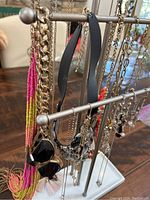 Jewelry stand showing assorted necklaces and bracelets hanging from multiple bars, including pink beaded necklace, black faceted necklace, and silver-tone chains with pendants and charms.