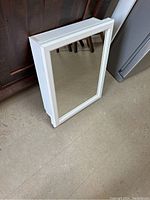 White floating mirrored cabinet shown standing against wood furniture and near metal appliance. Rectangular shape with framed mirror on front.