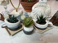 Photo showing three ceramic succulent planters with artificial green succulents, white Buddha statue, and two leaf-shaped candle holders with candles.