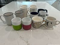 Photo showing fourteen assorted ceramic mugs on a countertop including plain, patterned, and color-blocked designs.