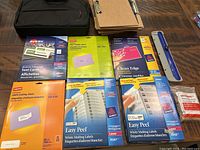 Photo showing black laptop bag, brown clipboards, packages of Avery and Staples mailing labels, name tags, rubber bands, and a blue ruler on wooden surface.